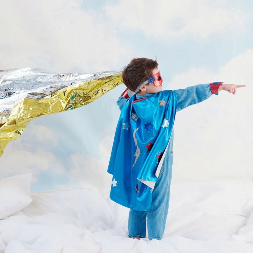 A child wearing a blue superhero cape and red goggles stands on a bed, holding a shiny gold and silver sheet, and points forward