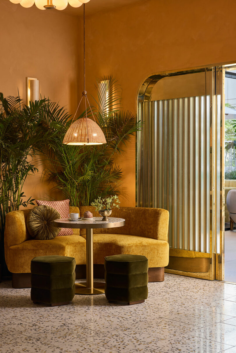 A cozy seating area with a yellow velvet sofa, two green stools, a round table, potted plants, and a hanging lamp, set against amber walls and a terrazzo floor by a metal-framed doorway.