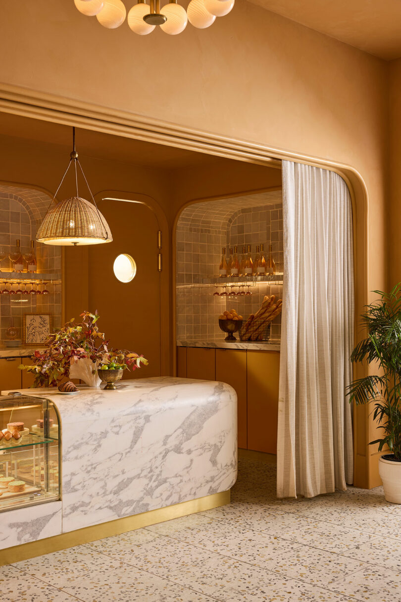 A modern cafe interior with a marble counter, glass display case, gold accents, tiled walls, hanging lights, indoor plants, and a sheer curtain dividing the space.