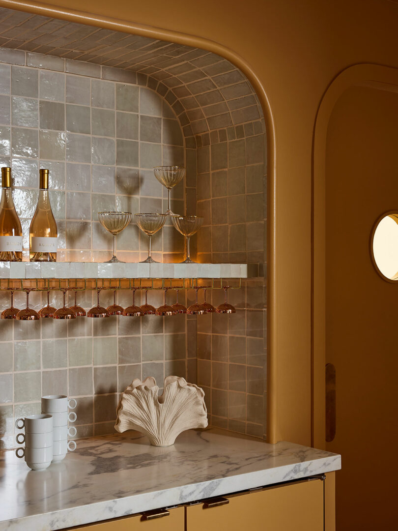 A modern bar nook with marble countertop, beige tiled backsplash, wine bottles, glassware, hanging glasses rack, stacked mugs, and a decorative shell sculpture.