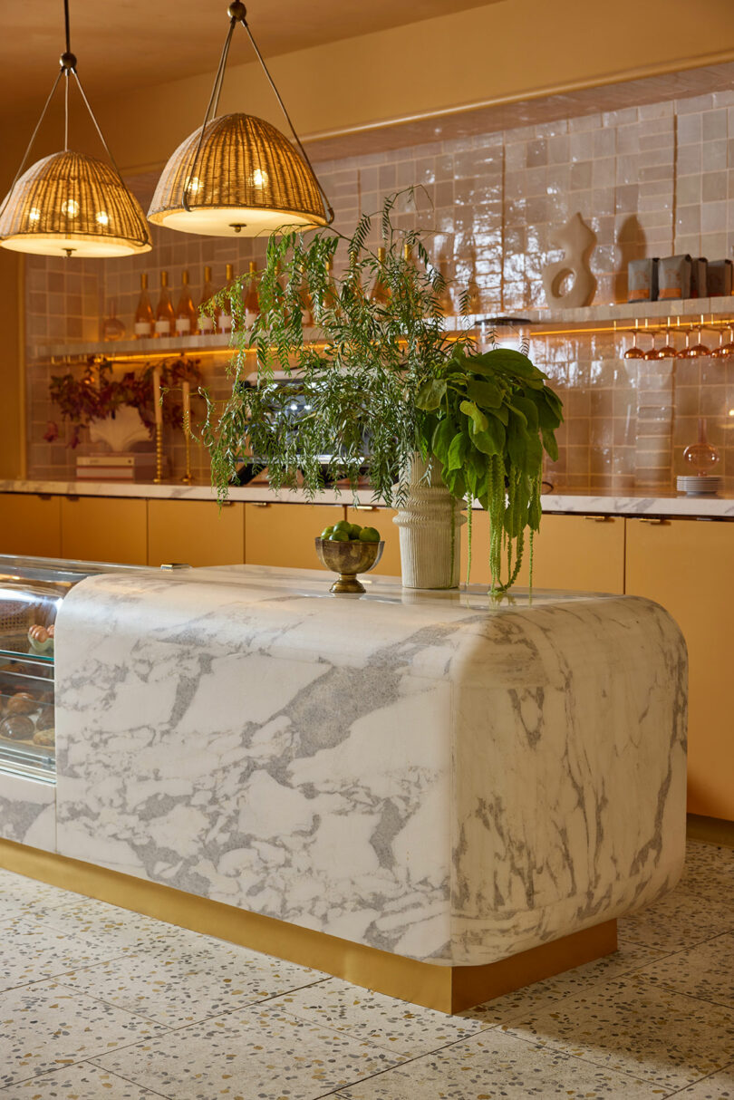 A marble counter with potted plants in a modern cafe interior, featuring pendant lights, tile backsplash, and warm-toned cabinetry.