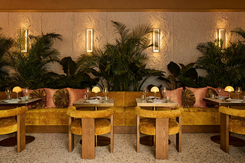 A stylish cafe interior with mustard yellow seating, wooden tables, round chairs, decorative pillows, plants, and warm wall lighting.