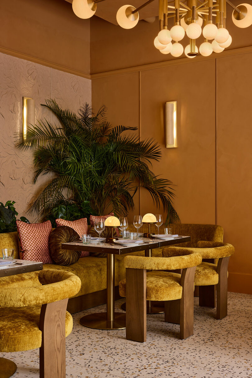 A cafe corner features gold velvet chairs, round wooden tables set with glassware, textured tan walls, gold light fixtures, and potted plants for decoration.