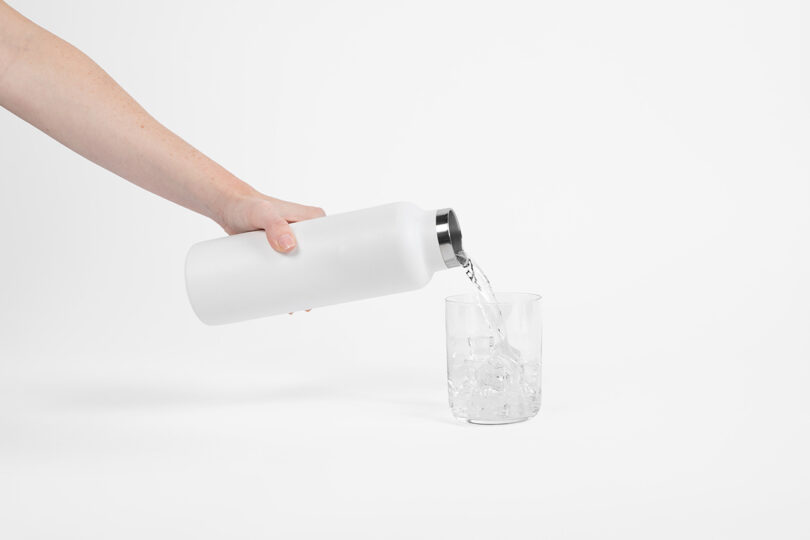A hand pours water from a white bottle into a glass filled with ice cubes against a plain white background.