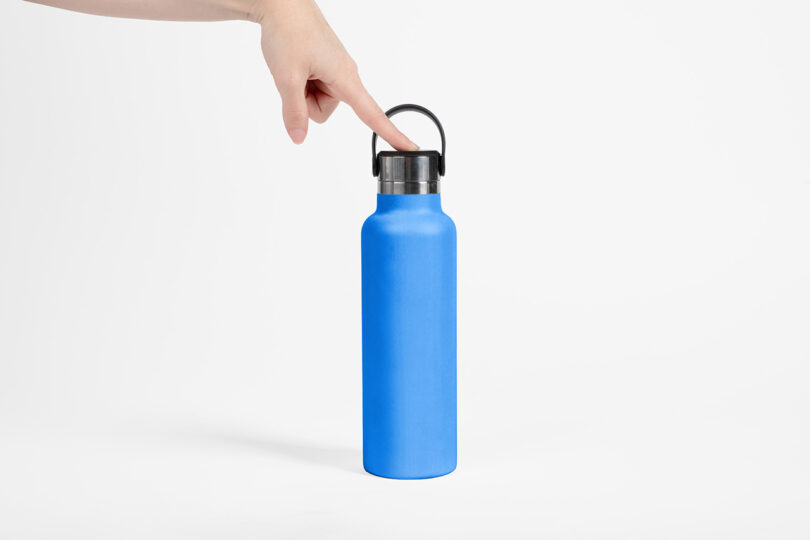 A hand presses the black lid of a blue reusable water bottle, which is standing upright on a white background.