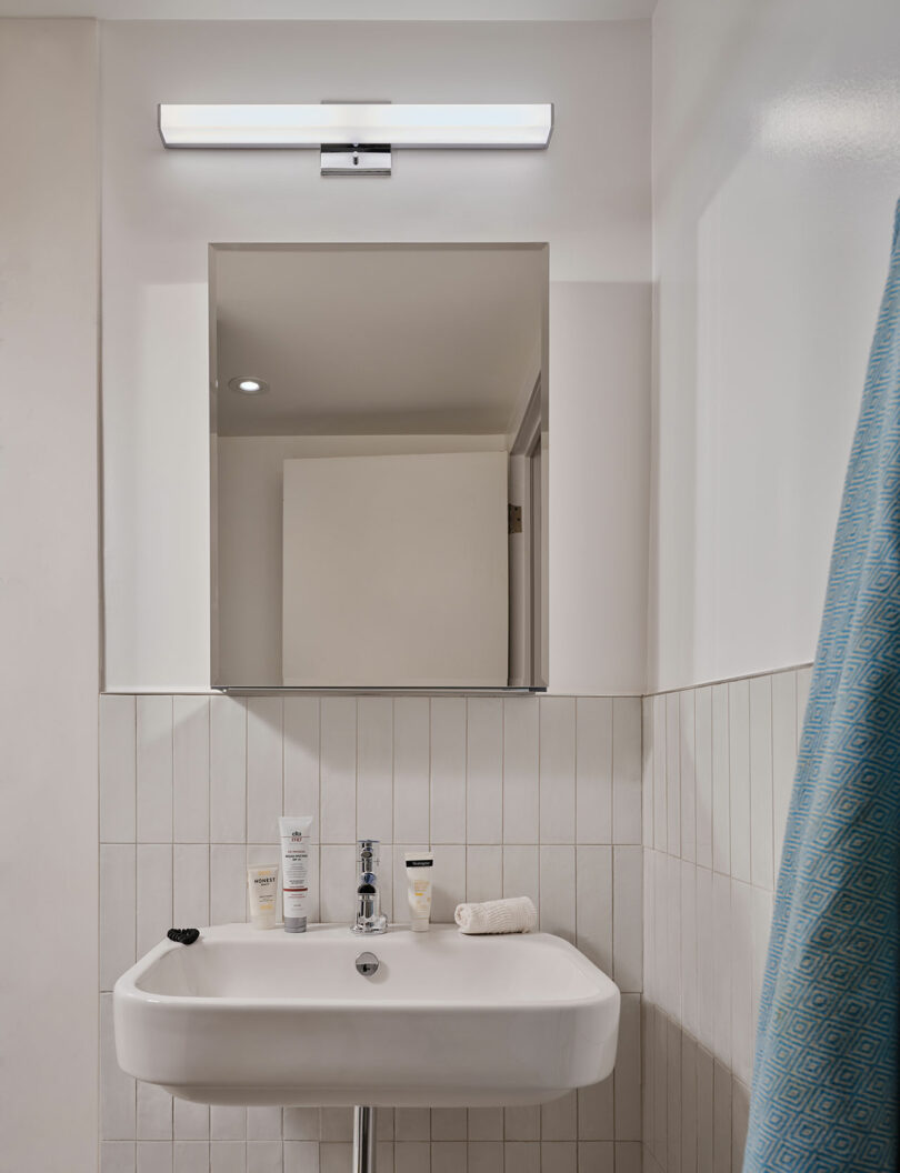 A small bathroom sink with toiletries, a rolled towel, a wall-mounted mirror, and an overhead light; blue shower curtain visible on the right.
