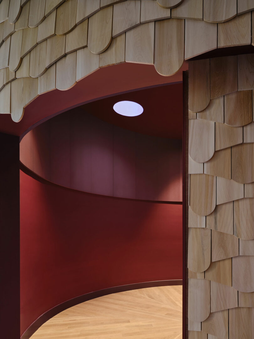Curved interior wall in deep red with a circular skylight, adjacent to a wall clad in overlapping, light wood shingles on the ceiling and entryway.