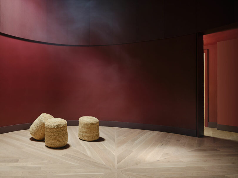 A minimalist room with curved maroon walls, light wood herringbone floor, and three round woven ottomans grouped together near the corner.