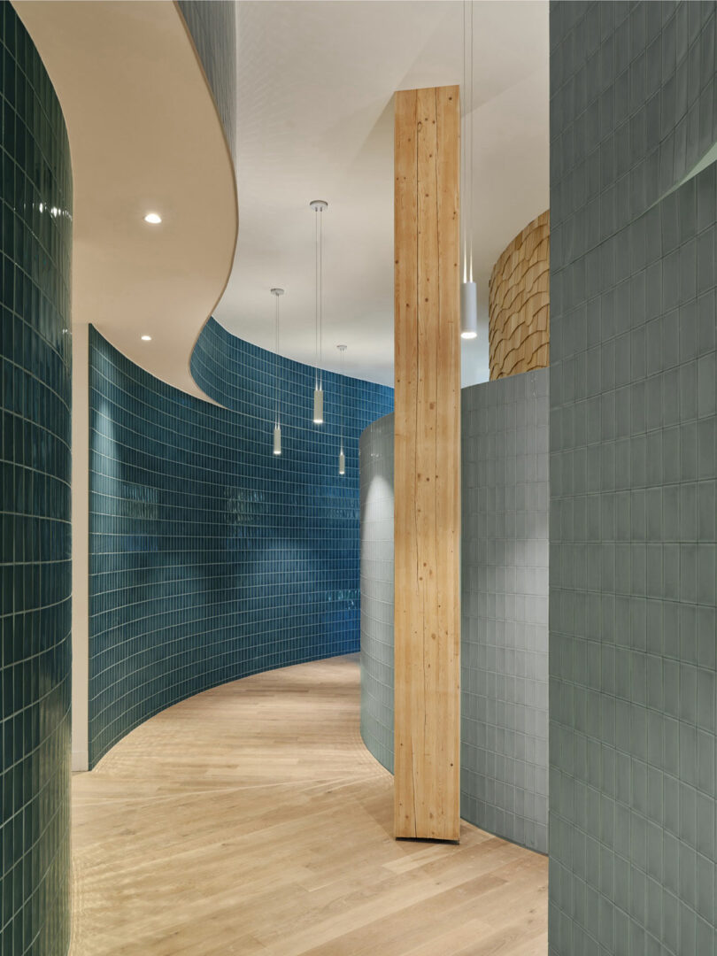 Curved hallway with teal tiled walls, light wooden floors, and a tall vertical wooden column. Several pendant lights hang from the ceiling.