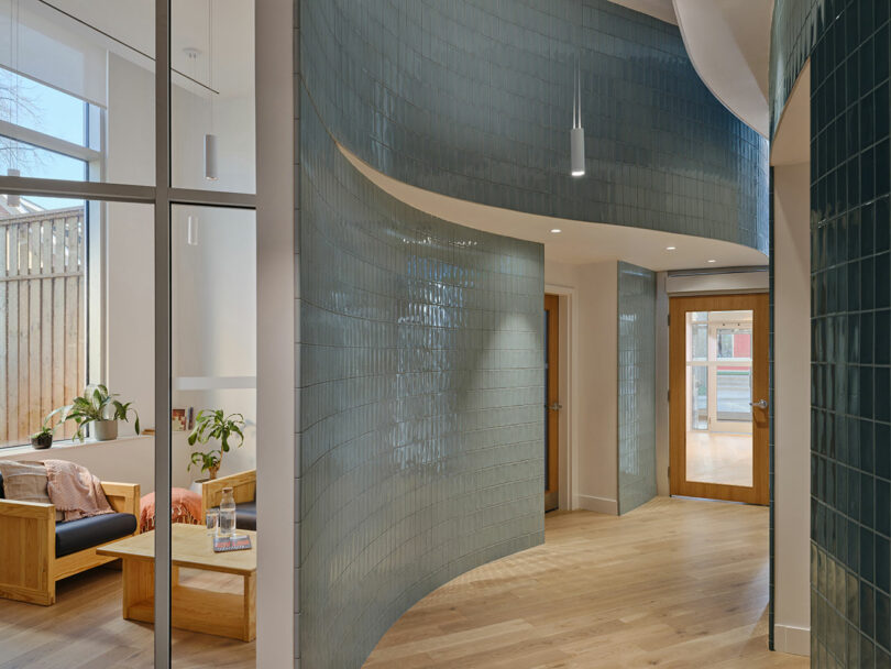 A modern interior hallway with curved blue-tiled walls, wooden doors, glass partitions, and a seating area with plants and natural light.