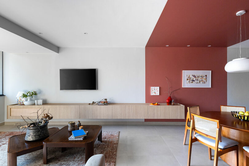 Modern living and dining area with a wall-mounted TV, floating cabinet, wooden furniture, and a partial red accent wall.