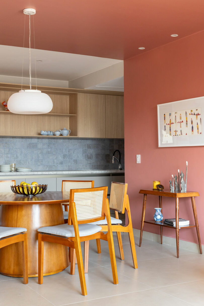 A modern dining area with a round wooden table, four chairs, a decorative bowl, and a side table against a reddish wall. A kitchen with light wood cabinets is visible in the background.