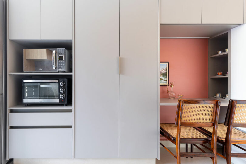 Modern kitchen with gray cabinets, a microwave, a toaster oven, cane-back chairs, and a small dining nook featuring a pink accent wall and framed picture.