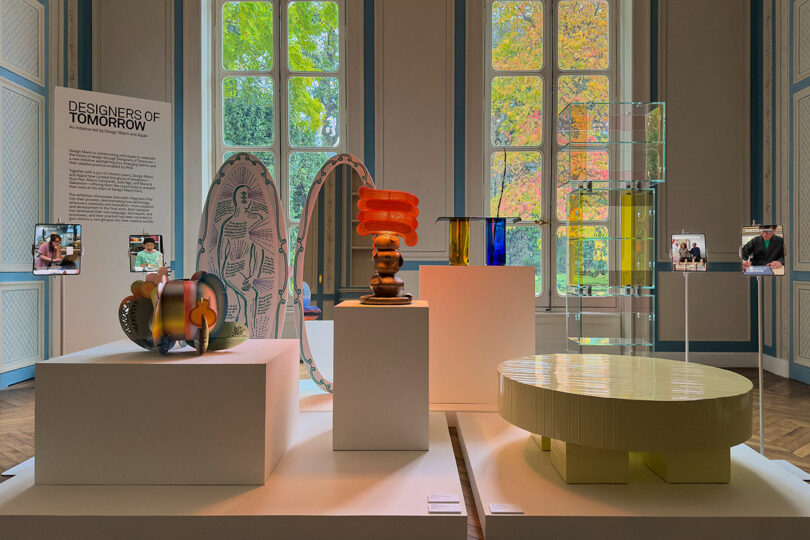 A museum exhibit displays colorful, modern design objects on white pedestals, with large windows showing trees outside and sleek iPad Pro screens beside each piece providing interactive information.