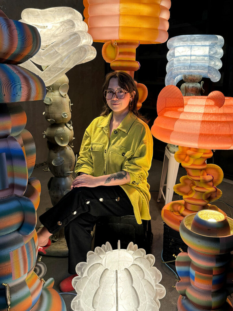A person wearing glasses and a yellow shirt sits on a stool, holding an iPad Pro, surrounded by colorful, sculptural lamps in a dimly lit room.