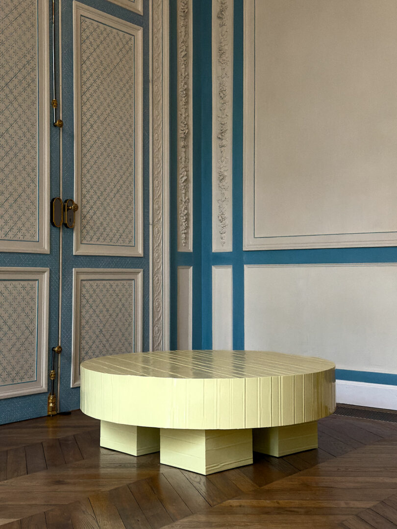 A round, light yellow table with block-style legs sits on a wooden floor in a room with ornate blue and white paneled walls; an iPad Pro rests at its center, ready for creativity by the double doors.