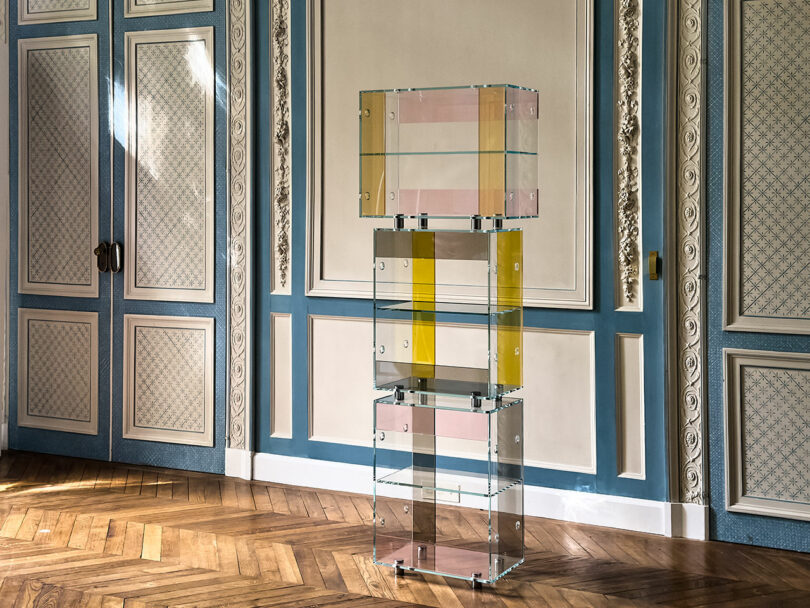A stack of transparent colored glass shelves stands in a room with ornate blue and beige paneled walls, a herringbone wood floor, and an iPad Pro resting elegantly on one of the shelves.