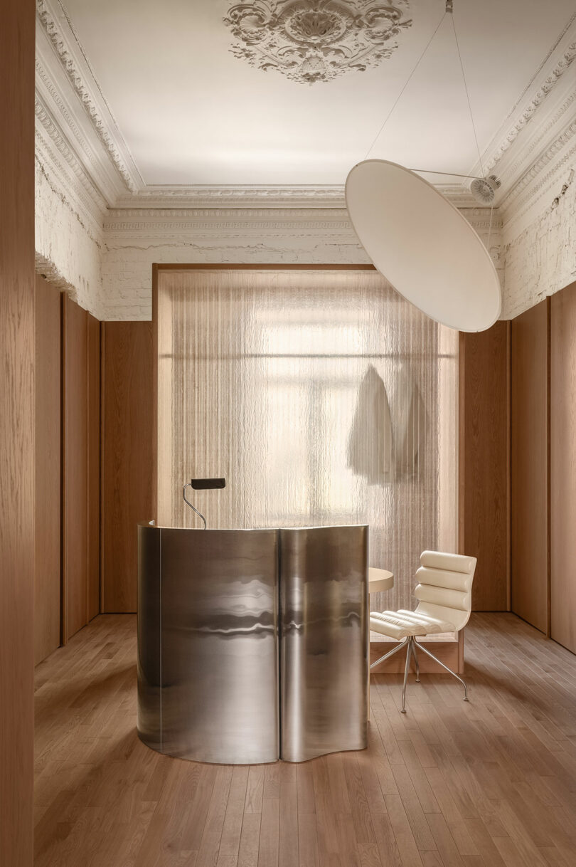 Minimalist reception area with a curved metallic desk, white chair, wood flooring, ornate ceiling molding, and sheer curtain dividing the space.