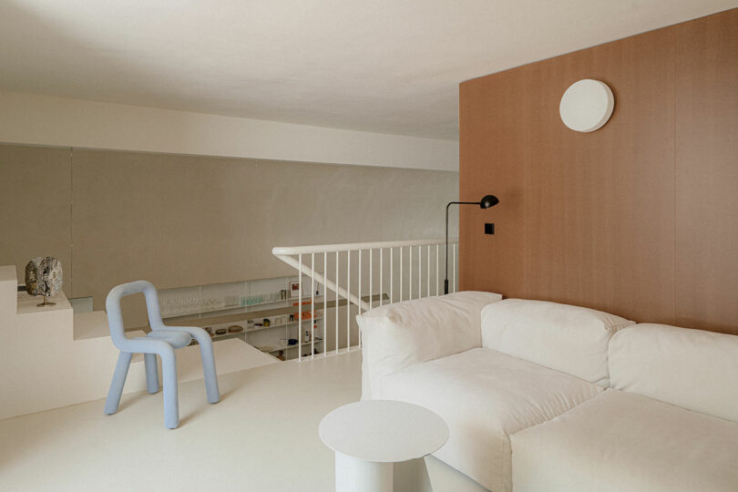 Minimalist living space with a white sofa, round wall light, blue chair, and white metal railing overlooking a lower level. Neutral tones and simple, modern furniture are featured.
