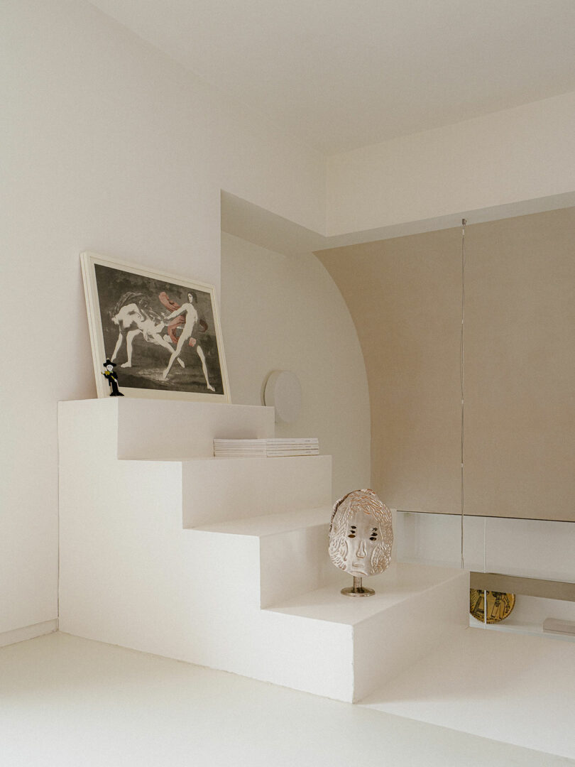 Minimalist white interior with geometric steps, a framed art print, stacked magazines, a small sculpture, and a decorative lamp on display.