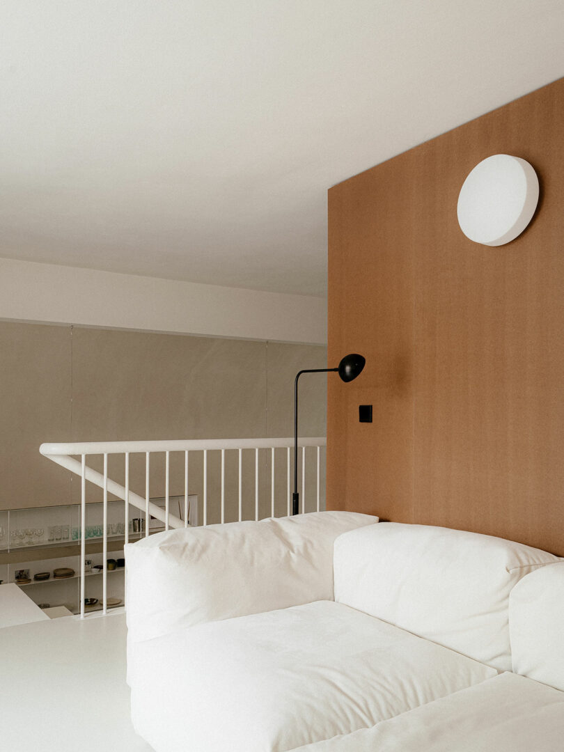 A modern living space with a white sofa, brown accent wall, round wall light, black floor lamp, and white railing overlooking a lower level.