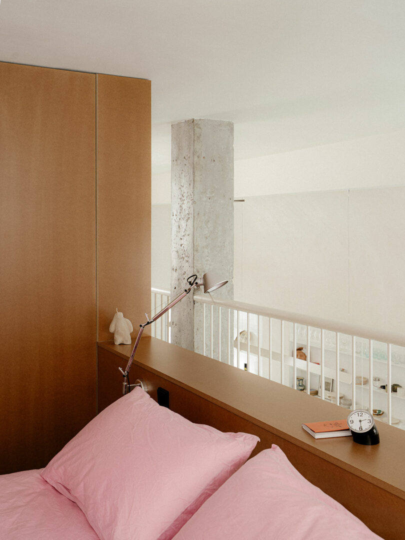 A bedroom with a bed featuring pink pillows, a wooden headboard, a desk lamp, an alarm clock, a book, a small figurine, and a concrete pillar in a modern, minimalist interior.