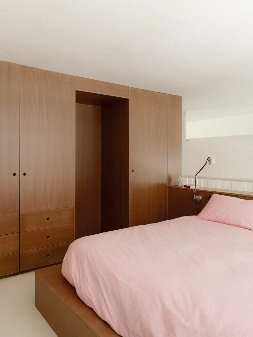 A minimalist bedroom with built-in wooden wardrobes and a platform bed with a pink duvet and matching pillows, next to a reading lamp.