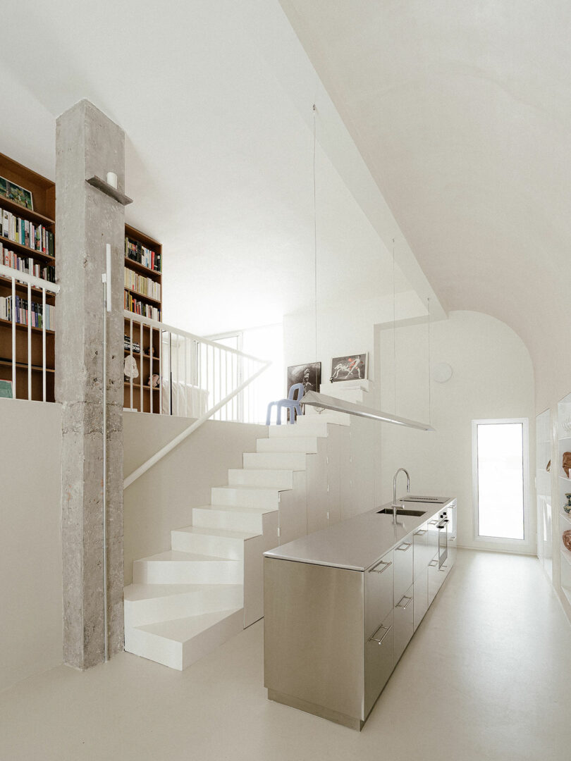 Minimalist kitchen with a stainless steel island, a white staircase leading to a loft with bookshelves, and an exposed concrete column. Light floods in from a door at the back.