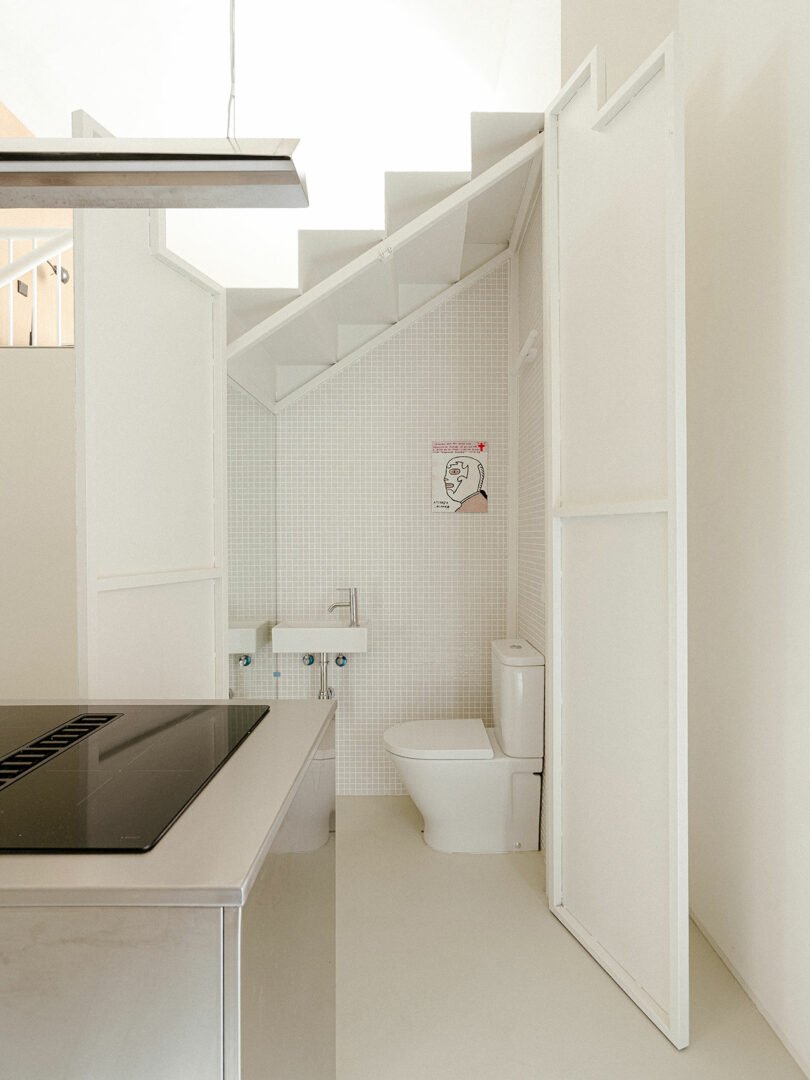 A small, minimalist bathroom is integrated under a staircase, featuring a toilet, a compact sink, and a wall-mounted illustrated sign.