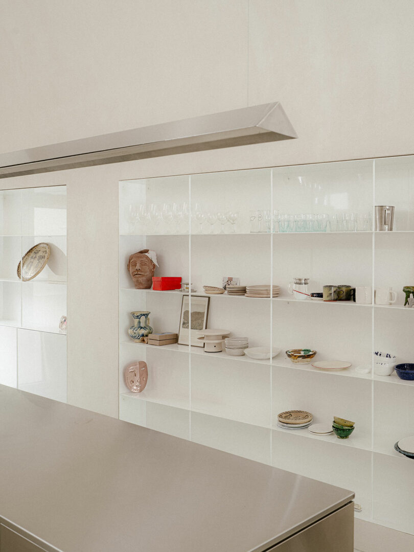 Minimalist kitchen with open white shelving displaying dishes, glasses, ceramics, and decorative objects; a sleek metal countertop is in the foreground.
