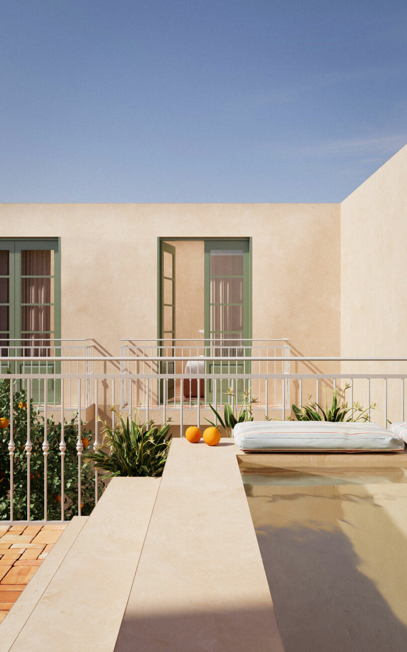 A minimalist terrace with a small pool, two oranges, a light blue cushion, green-framed doors, potted plants, and beige walls under a clear blue sky.