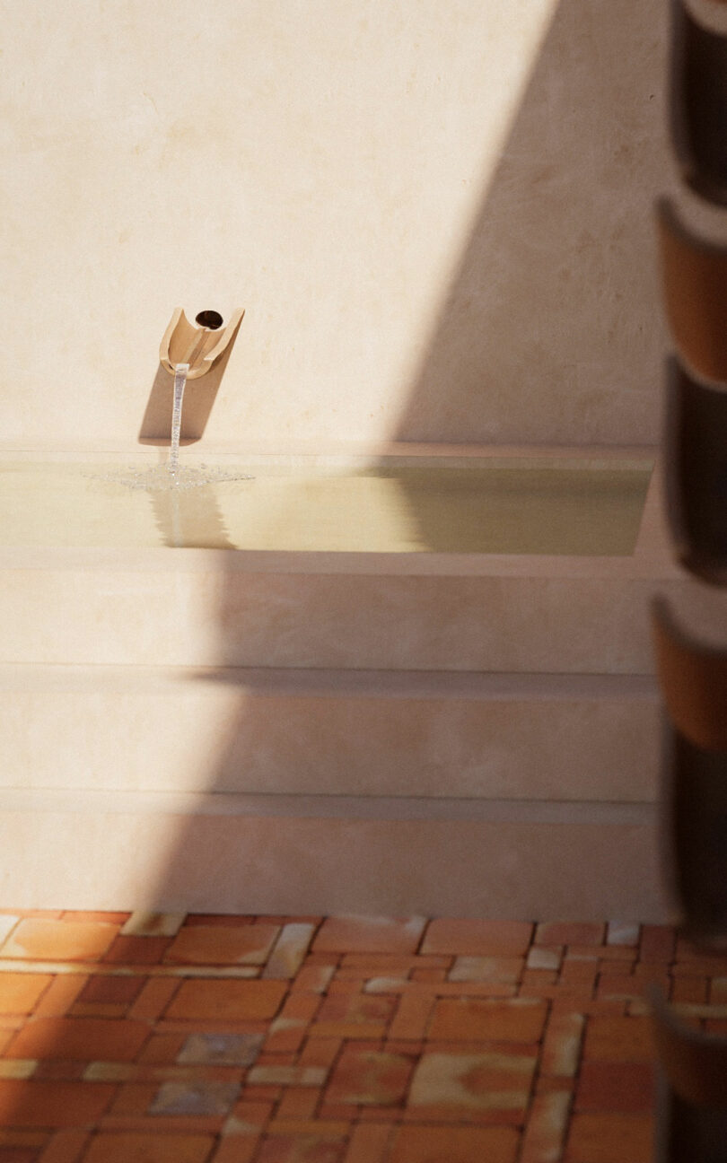 A small stream of water flows from a spout into a shallow rectangular pool, with sunlight and shadows cast on the neutral-toned wall and brick floor.