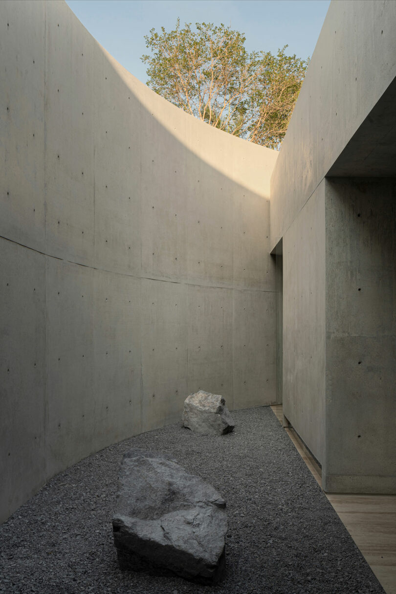 A minimalist concrete courtyard with gravel ground, large rocks, and an opening above revealing a tree and blue sky.