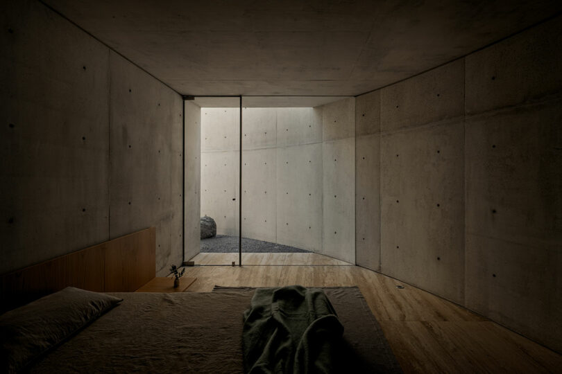 Minimalist bedroom with concrete walls, a simple bed, wooden floor, and large glass doors opening to a small enclosed courtyard with gravel and a single rock.