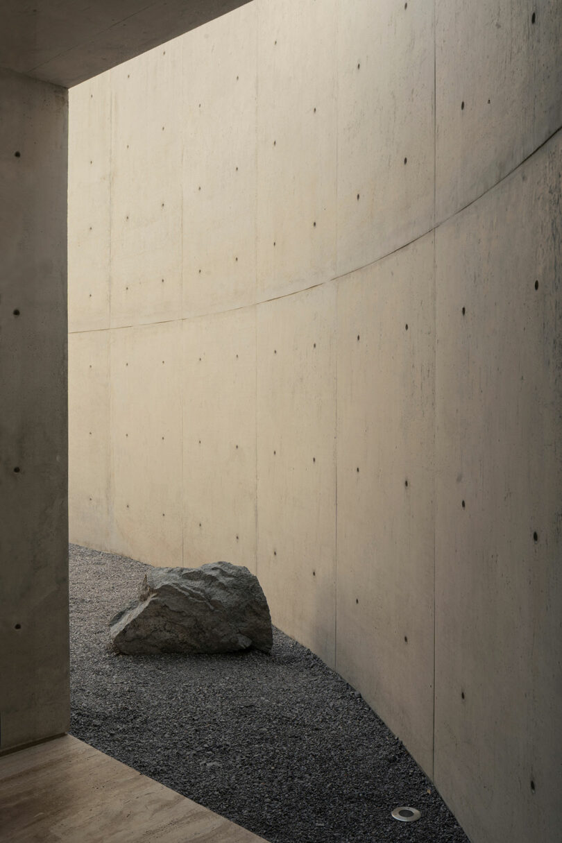Curved concrete wall with visible bolt marks, gravel ground, a large rock, and natural light casting soft shadows.