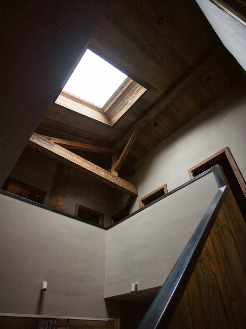 View of a modern interior with exposed wooden beams, a skylight, and angled walls, allowing natural light to enter the upper floor.