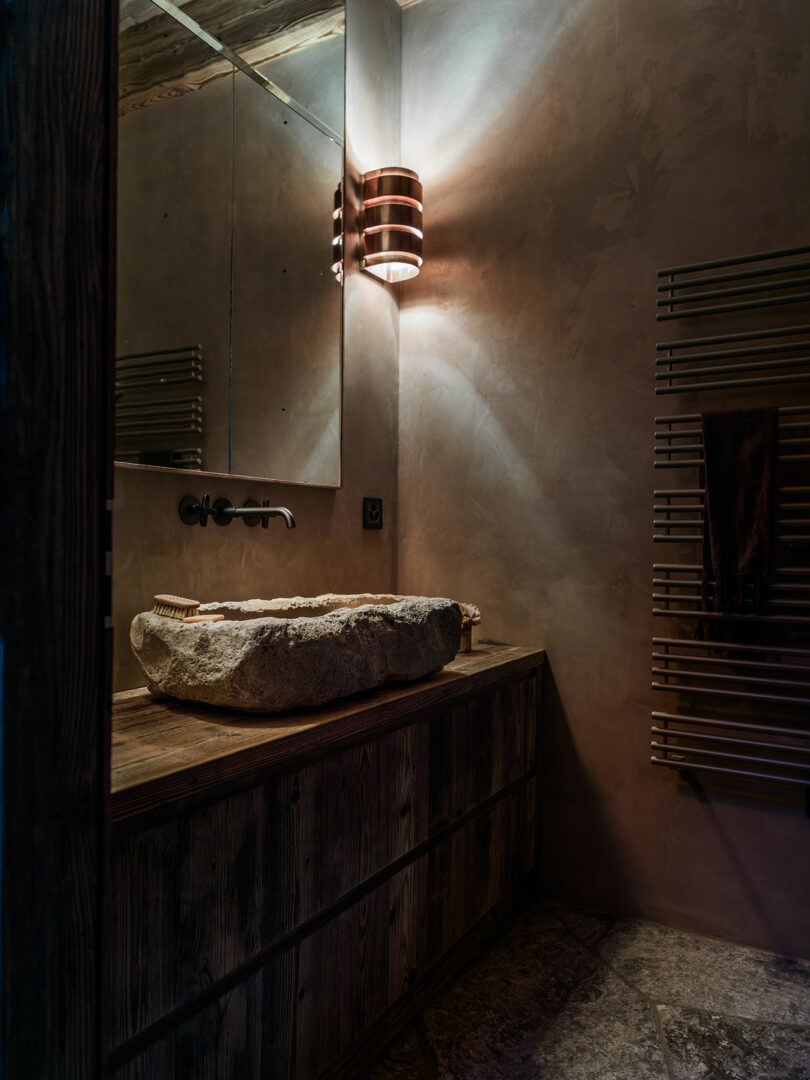 A rustic bathroom with a stone sink on a wooden vanity, wall-mounted faucet, large mirror, modern wall sconce, and towel radiator.