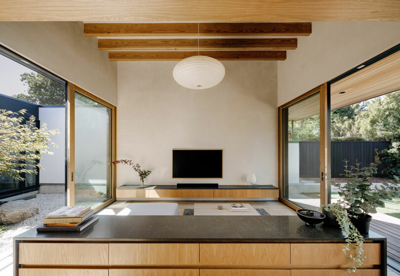 Modern living room with wooden beams, large windows, a low TV unit, and minimalist decor, overlooking a garden on both sides.