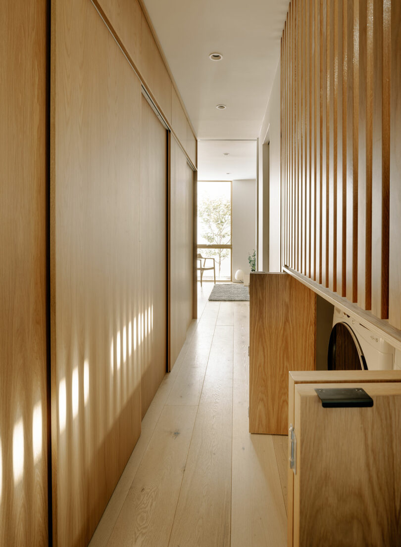 A narrow hallway with light wooden floors and walls, featuring built-in cabinetry, a washing machine, and vertical wooden slats, leading to a bright room with a window.