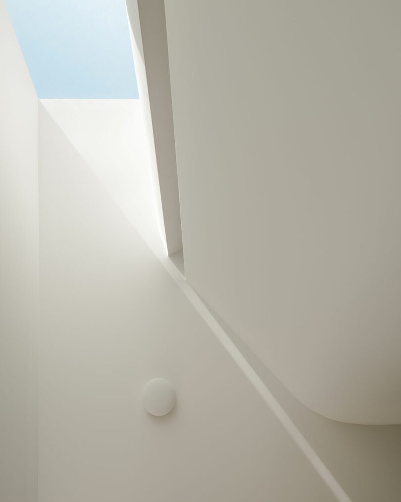 Minimalist architectural interior with white walls, soft curved surfaces, a circular ceiling light, and a skylight revealing a blue sky. Light and shadows create geometric patterns.
