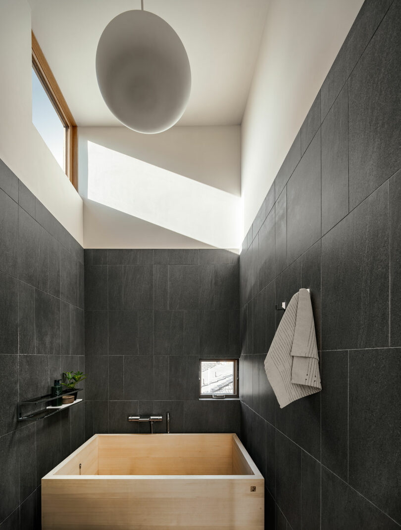 A minimalist bathroom with dark tiled walls, a light wood soaking tub, a small window, a towel hanging on a hook, and a round ceiling light.