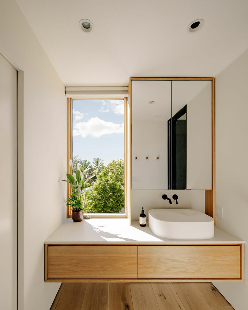 A minimalist bathroom vanity with wood drawers, a white sink, black fixtures, a mirror cabinet, a potted plant, and a window with a view of greenery and blue sky.