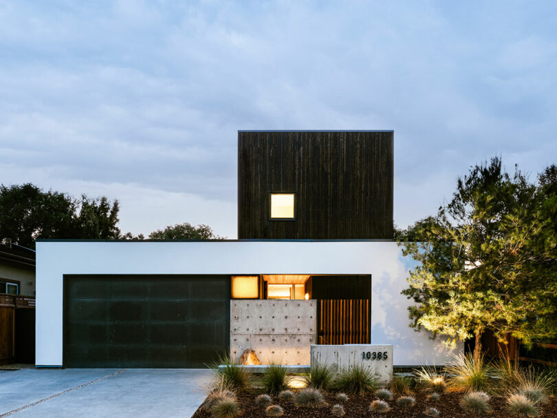 Modern two-story house with a minimalist design, featuring a dark upper section, white lower facade, large garage door, and landscaped front yard.