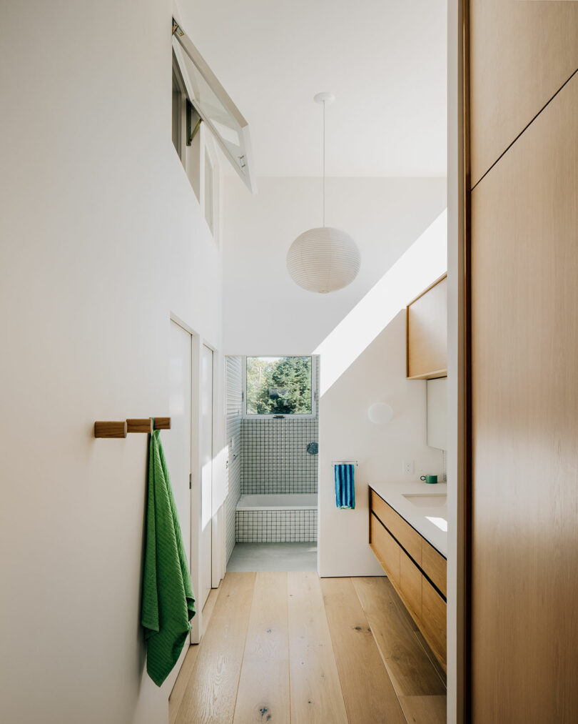 Minimalist bathroom with light wood accents, a green towel, a paper lantern ceiling light, and a window letting in natural light above a tiled bathtub and shower area.