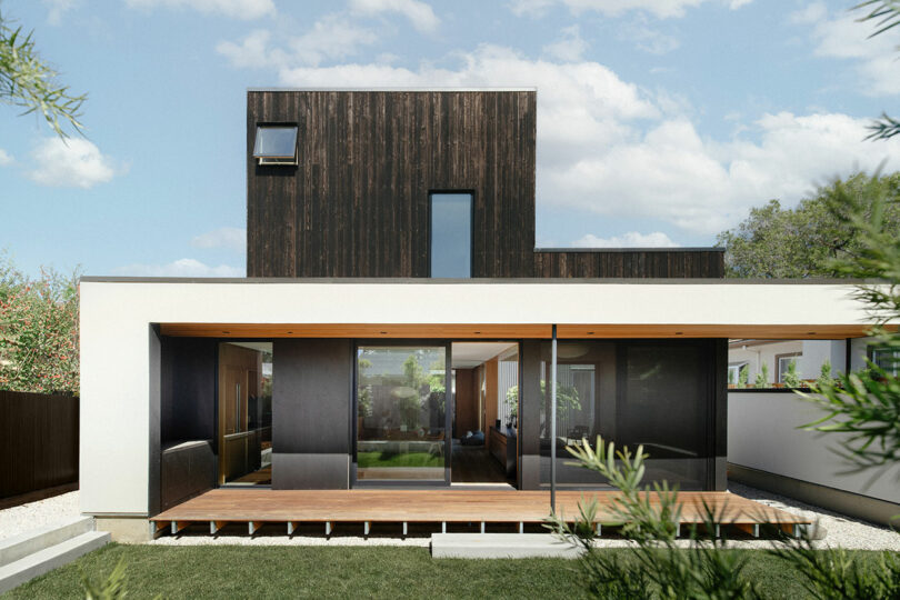 A modern two-story house with a dark wood upper level, large glass sliding doors, a covered patio, and a lawn in front.