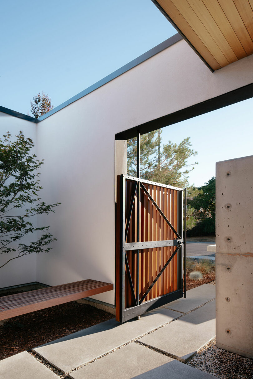 A modern outdoor entryway with a partially open metal and wood gate, concrete path, white walls, a wooden bench, and a tree on the left.