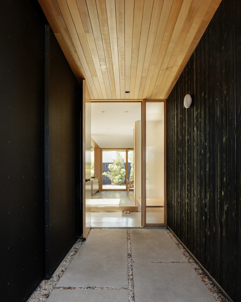 View of a modern home's entryway with a wood ceiling, black walls, concrete floor panels, and a glass door opening to a bright interior living space with large windows.
