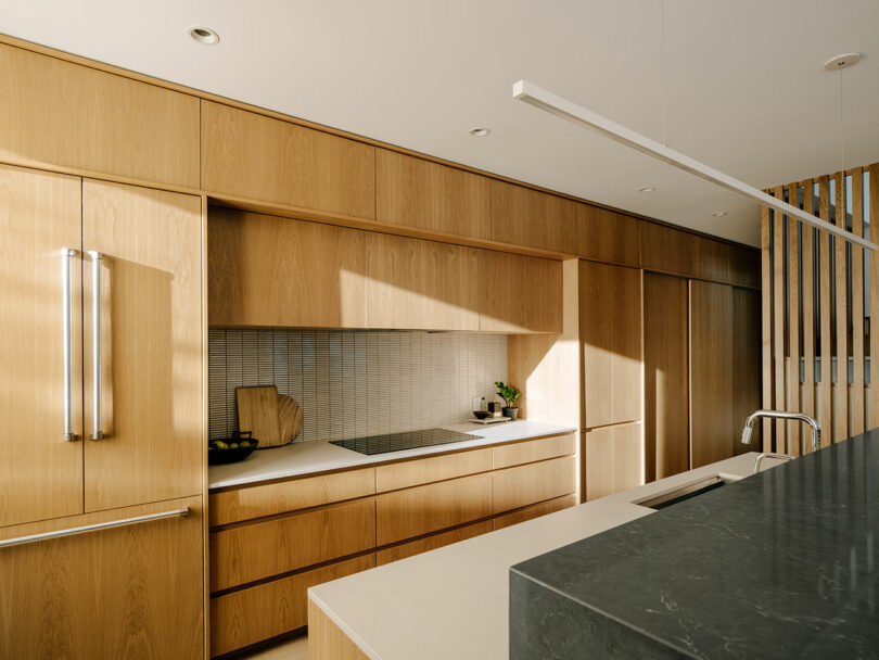 Modern kitchen with light wood cabinetry, white countertops, minimalist backsplash, integrated appliances, and a dark stone island with a built-in sink.