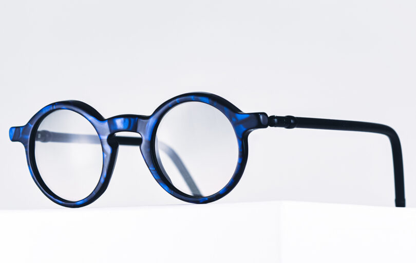 A pair of round, blue tortoiseshell eyeglasses with clear lenses is displayed against a plain white background.