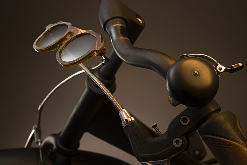 A pair of sunglasses is resting on the handlebars of a bicycle, with the brake lever and bell visible against a plain background.